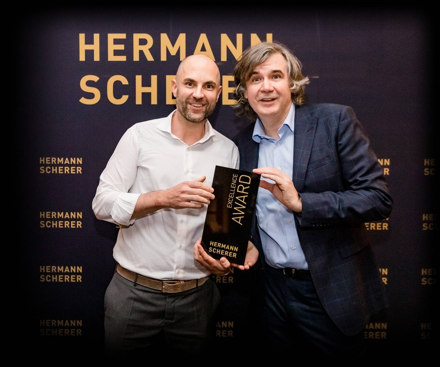 Dr. med. Niels Freitag und Hermann Scherer halten gemeinsam die Trophäe des Excellence Award, dahinter eine Wand mit dem Schriftzug Hermann Scherer